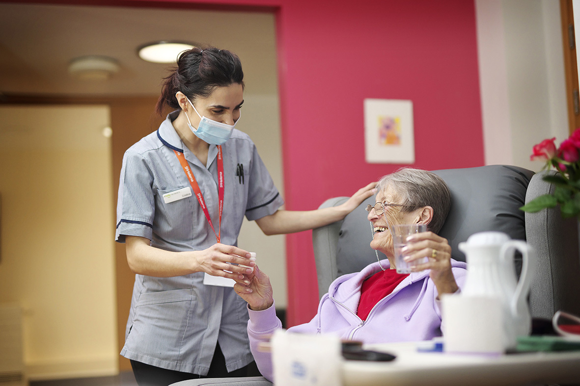 A nurse stands next to a patient who is sitting in and armchair and smiling