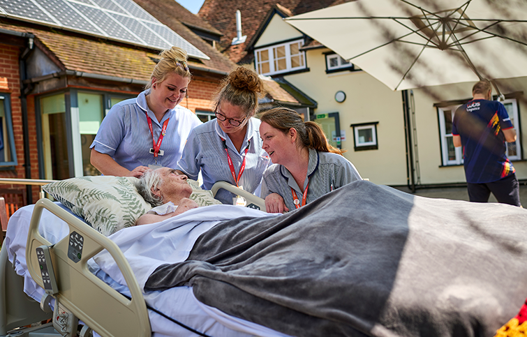 Three St Helena staff smiling at a patient laying in a bed in the hospice garden