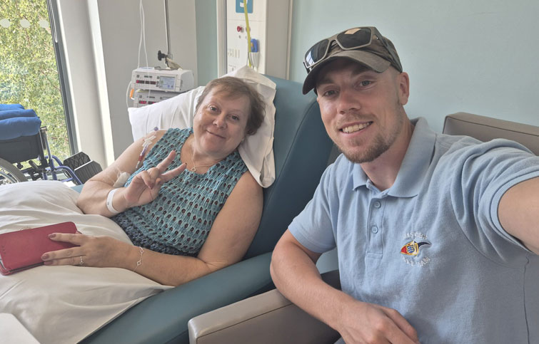 Wendy is in a chair having chemo and her son Alex is sitting next to her, both are smiling at the camera