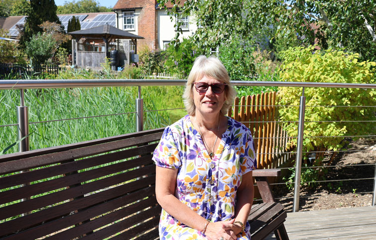 Sharon sitting on a garden bench with St Helena Hospice behind her