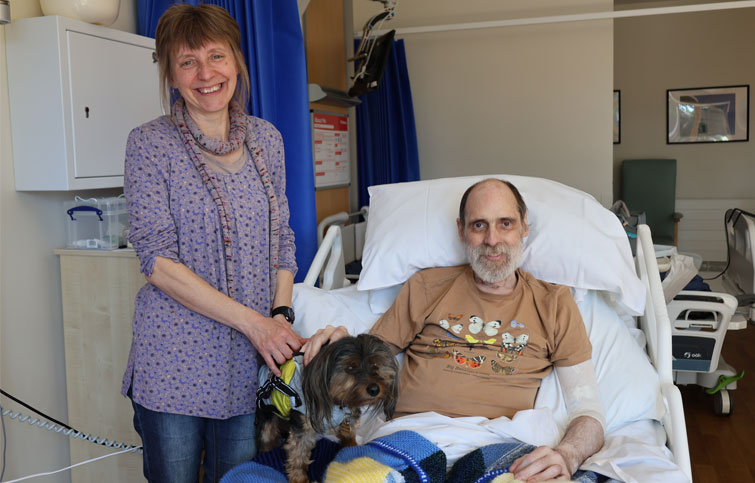 Claire is standing next to Bradley who is in his St Helena Hospice bed with their little dog beside him on the bed, they are smiling at the camera