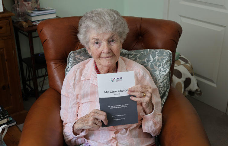 Kay is sitting in a chair looking at the camera and she is hold a My Care Choices Record